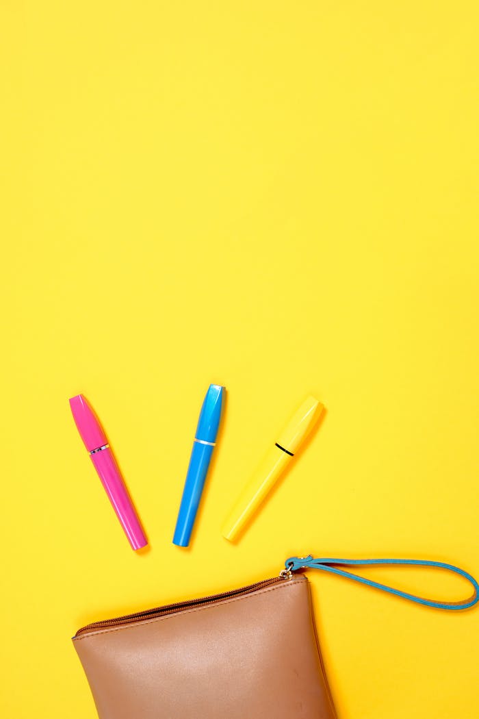 Flat lay of colorful mascara tubes and cosmetic pouch on a bright yellow backdrop.